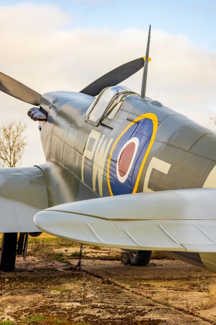 A vintage World War II fighter aircraft, likely a Spitfire, is displayed outdoors on a grassy area under a partly cloudy sky. The airplane's fuselage features a shiny metallic surface with distinctive