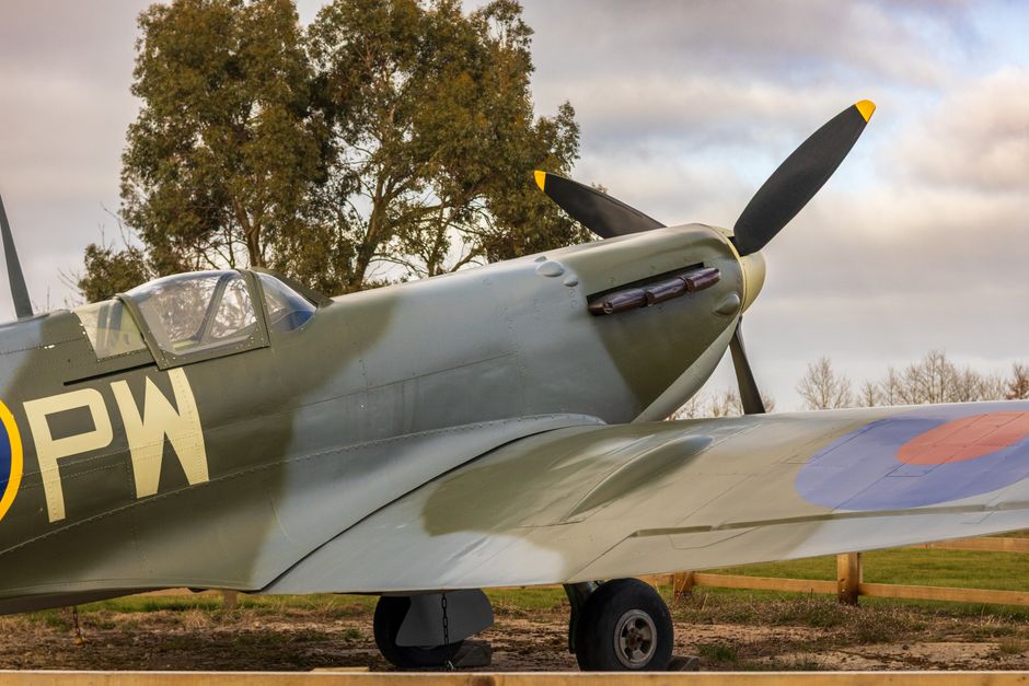 A vintage World War II fighter aircraft, likely a Spitfire, is displayed outdoors on a grassy area under a partly cloudy sky. The airplane's fuselage features a shiny metallic surface with distinctive